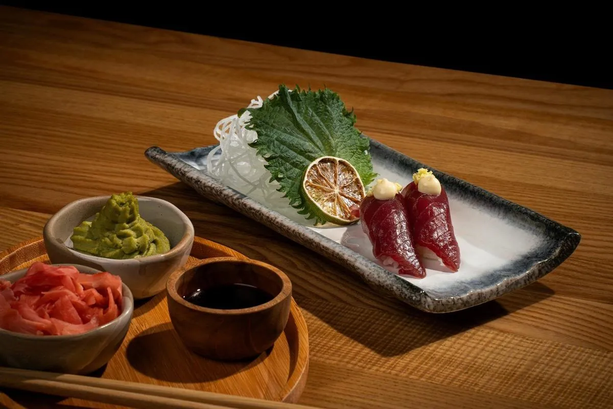 Two pieces of tuna nigiri — raw tuna draped over compressed rice — on a ceramic plate with shiso, daikon and citrus
